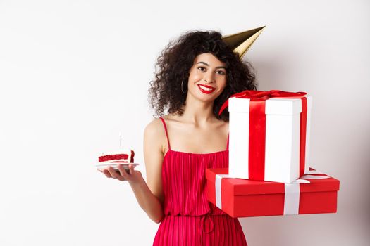 Holidays. Happy elegant woman in party hat and red dress, holding birthday cake and gifts, celebrating b-day, standing over white background.