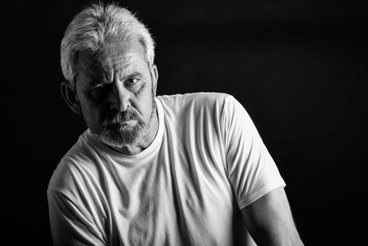 Portrait of a serious mature man looking at camera. Senior male with white hair and beard wearing casual clothes isolated on black background. Studio shot in black and white.