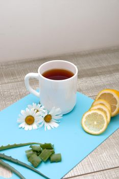 proper nutrition, white cup with chamomile tea on a blue cutting board with chamomile flowers, sliced lemon, aloe and polka dot fabric on a gray wooden table