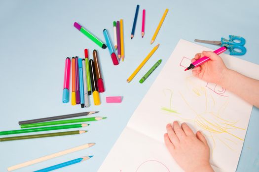 a small child draws with a pink felt-tip pen in an album, copy space, top view, blue background, pencils, wax crayons, scissors and the child's hands on the table