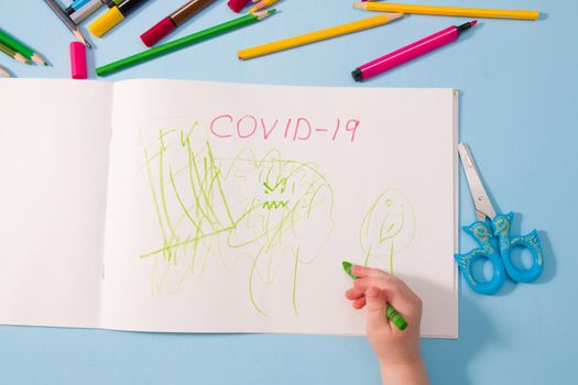 a small child draws green wax crayon in an album coronavirus, pencils on a blue background, top view, copy space, activities with children at home during quarantine