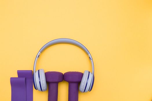 Fitness and healthy active lifestyle background concep,blue and gray colored wireless headphones, elastic band expanders and purple dumbbells on a yellow background, copy space, top view