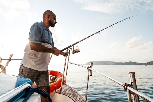 Young african american man standing with fishing rod on a sailboat fishing in open sea on sunset, close up