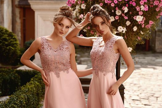 Beautiful blonde and brunette bridesmaids with luxury hairdo in gorgeous elegant stylish pale pink floor length chiffon gown dress decorated with sequins sparkles and rhinestones on flowers background. Wedding day in old beautiful European city.