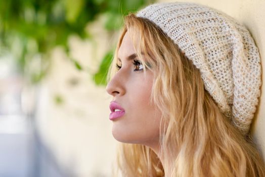 Close-up profile of blonde young woman with curly hair. Girl wearing sweater and wool cap in urban background. Female with beautiful lips painted in pink