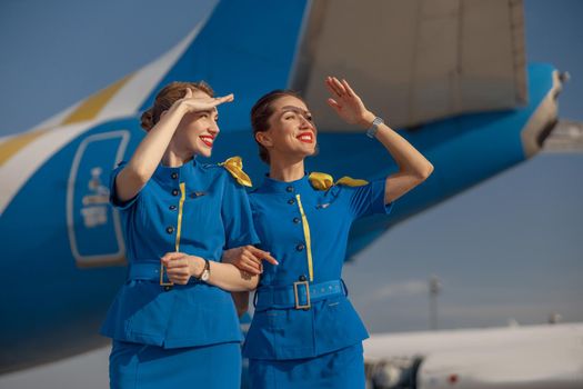 Portrait of two cheerful air stewardesses in bright blue uniform walking outdoors in front of passenger aircraft on a sunny day. Occupation concept