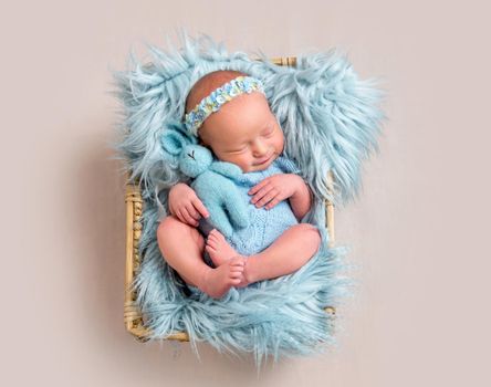 Happy newborn girl lying on her back and cuddling her toy. Little girl wearing blue headband and blue bodysuit lying on blue flokati rug and smiles in sleep while hugging her hare toy