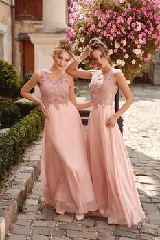 Beautiful blonde and brunette bridesmaids with luxury hairdo in gorgeous elegant stylish pale pink floor length chiffon gown dress decorated with sequins sparkles and rhinestones on flowers background. Wedding day in old beautiful European city.