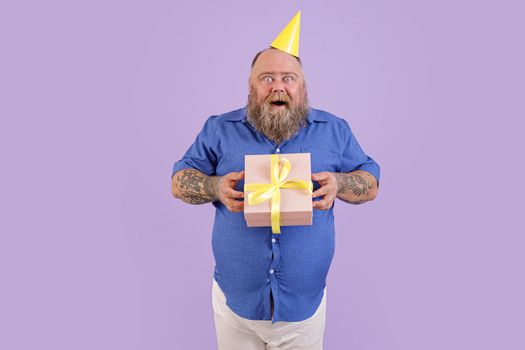 Excited mature plump man wearing tight blue shirt and party hat holds present posing on purple background in studio