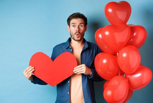 Valentines day concept. Boyfriend in love looking amazed at camera, holding red paper heart and standing with romantic balloon, blue background.