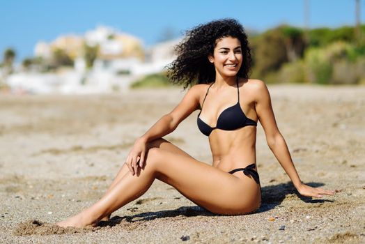 Young arabic woman with beautiful body in swimwear sitting on the beach sand. Smiling female with curly long hairstyle wearing black bikini.