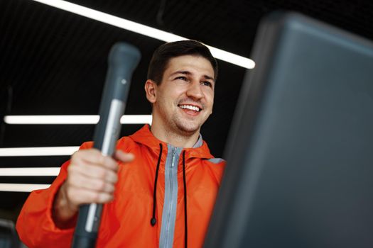 Portrait of a young man in orange windbreaker workout on a fitness machine at a gym.