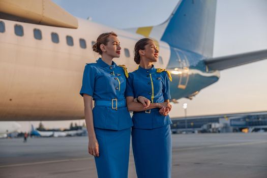 Two beautiful air hostesses in blue uniform smiling away, standing in front of a big passenger airplane in airport at sunset. Aircrew, occupation concept