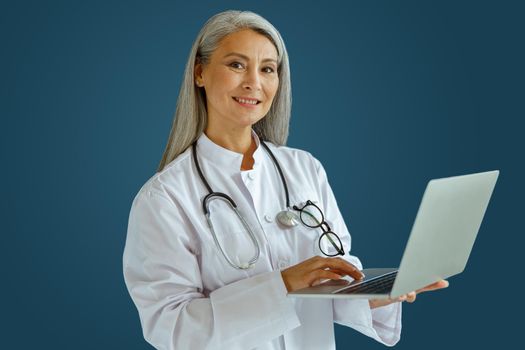 Smiling mature woman doctor in white coat works on laptop standing on blue background in studio. Telemedicine patient service
