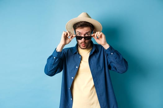 Portrait of tourist takes-off sunglasses and saying wow, staring impressed at camera, checking out cool thing, standing in summer hat on blue background.