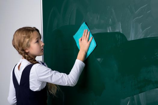 Schoolgirl wiping board with rag in classroom. Primary school student in uniform standing near of blackboard and erasing it. Back to school, education concept
