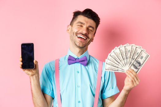 E-commerce and shopping concept. Rich and happy guy laughing, showing money in cash and empty smartphone screen, standing on pink background.
