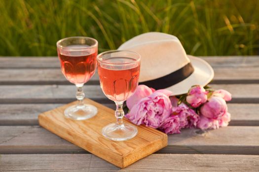 two glasses with rose wine and pink peony bouquet. delicious refreshing drink, huge blossoming flowers and mens hat on wooden pier at sunset