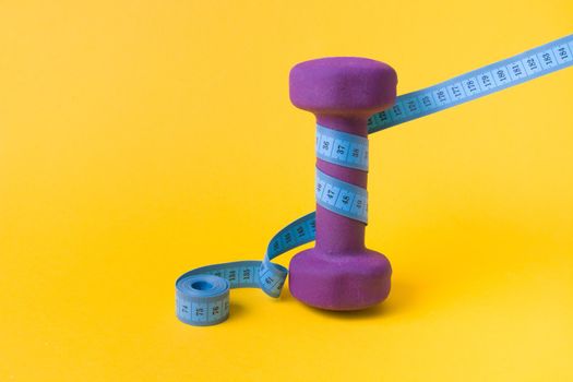 1.5 kilogram purple dumbbell and blue measuring tape on a yellow background, a dumbbell wrapped in a folded tape copy space, weight loss concept