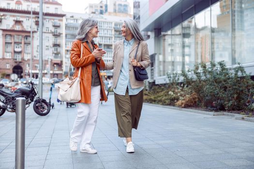 Cheerful senior woman with long haired companion walk on large city street on nice autumn day. Friends spend time together