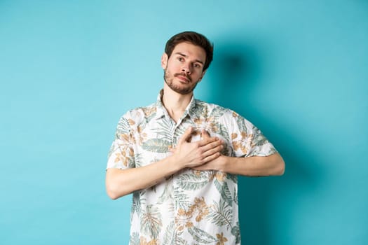 Handsome young man in summer shirt, holding hands on heart and looking thankful, being grateful, standing on blue background.