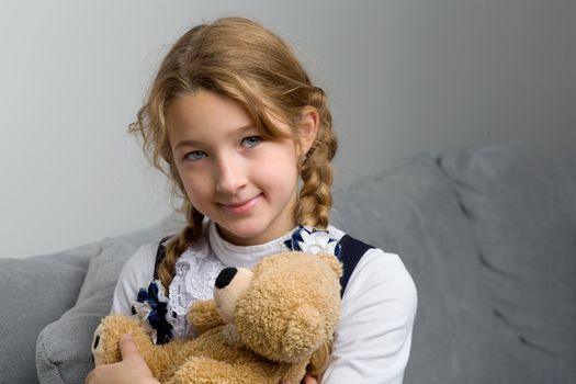 Lovely blonde girl sitting on comfortable sofa hugging teddy bear. Portrait of cute long haired girl wearing warm knitted dress. Adorable happy child resting on couch with favorite stuffed toy