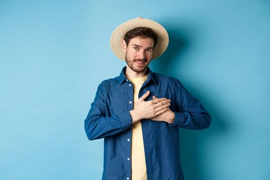 Handsome traveler in straw hat, holding hands on heart and smiling, showing thank you gesture, keep memories about vacation in soul, standing on blue background.