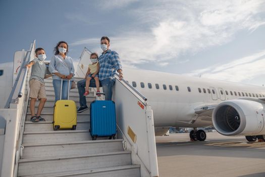 Happy family in protective masks, parents with two little children standing on airstair, boarding the plane in the daytime. People, traveling, vacation concept