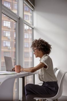 Side view of smiling young lady wearing glasses while working with laptop and typing in the office