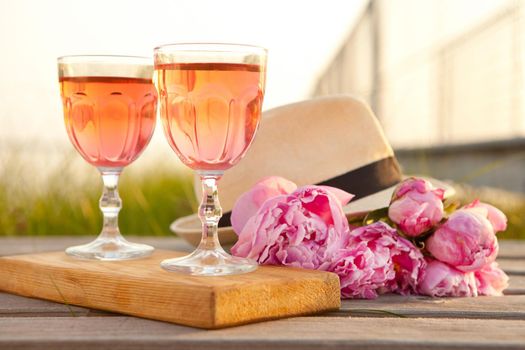 two glasses with rose wine and pink peony bouquet. delicious refreshing drink, huge blossoming flowers and mens hat on wooden pier at sunset