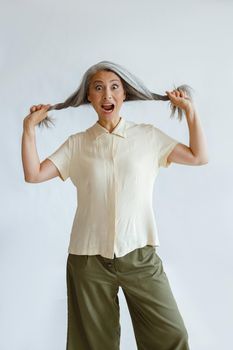 Funny shocked Asian woman in stylish clothes holds long hoary hair locks on light grey background in studio. Mature beauty lifestyle