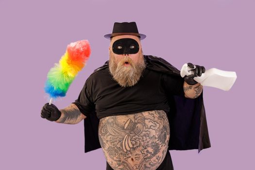 Surprised man with overweight in carnival Zorro suit with large bare tummy holds dust brush and spray bottle on purple background in studio
