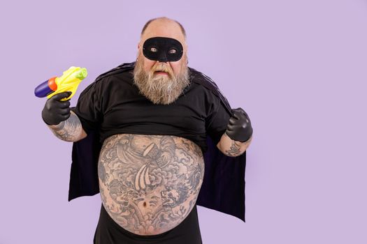 Funny evil obese man wearing carnival costume with cape and mask holds toy blaster posing on purple background in studio