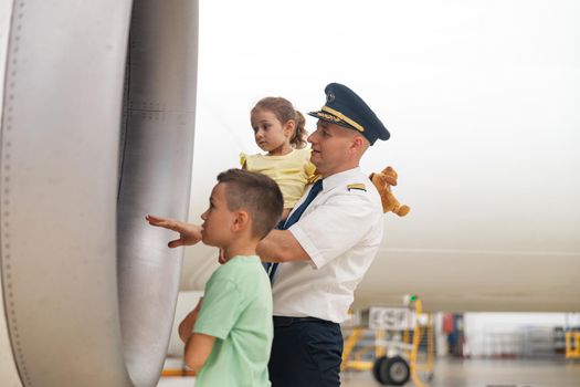 Pilot in uniform showing parts of airplane to two little kids who came to excursion at the aircraft hangar. Aircraft, excursion, childhood concept