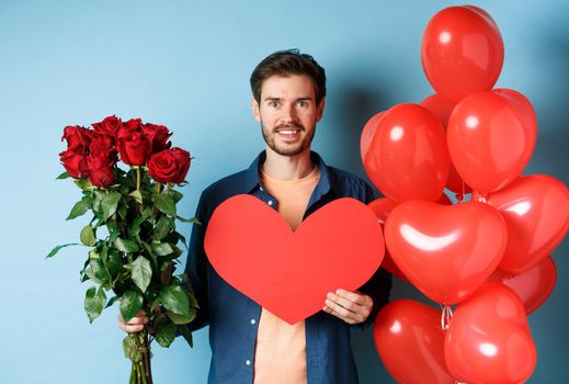 Valentines day romance. Boyfriend with bouquet of red roses and heart balloons smiling, bring presents for lover on valentine date, standing over blue background.