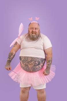 Funny brutal middle aged plus size man wearing fairy costume with magic stick and wings stands on purple background in studio