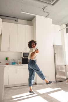 Full-length photo of happy multiethnic female wearing wireless headphones and dancing on the kitchen while holding smartphone
