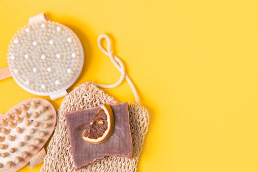 dry anti-cellulite massage brush, knitted washcloth, homemade cocoa soap, dried orange slices and a wooden body massager on a yellow background, top view, copy space, flat lay