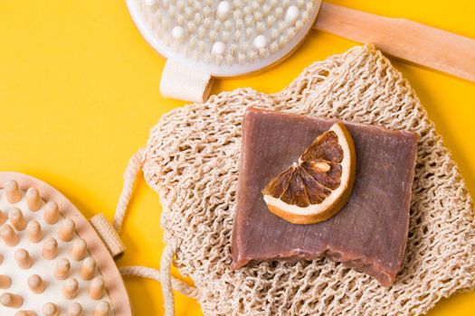 dry anti-cellulite massage brush, knitted washcloth, homemade cocoa soap, dried orange slices and a wooden body massager on a yellow background, top view, copy space, flat lay