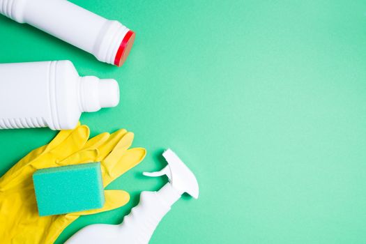 yellow rubber gloves, green sponges for washing and several different types of white plastic bottles without labels for detergents on a green background top view copy space