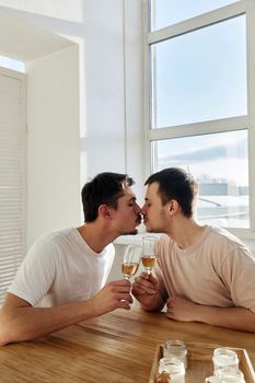 Young boyfriends clinking wineglasses and kissing each other while sitting at table during romantic date on weekend day at home