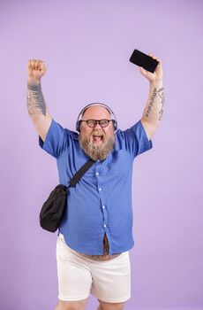 Excited middle aged fat man with headphones and crossbody bag raises up hands and cellphone with blank screen posing on purple background in studio