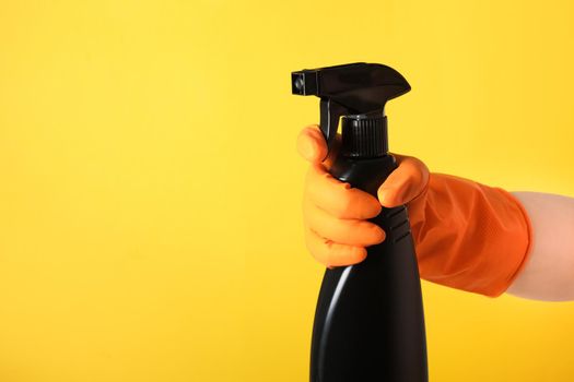 A hand in a orange glove holds a black spray bottle of cleaning fluid on a yellow background copy space