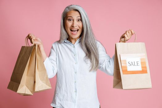 Excited mature Asian woman holds paper shopping bags with colorful Sale sign standing on pink background in studio