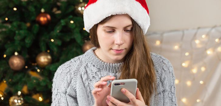 Online christmas shopping. Cute beautiful attractive woman browsing through her phone in search of something while wearing santa hat and gray sweater. quarantine shopping concept