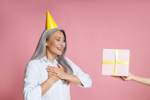 Happy middle aged Asian lady with long grey hair and yellow party hat looks at gift box on friend hand on pink background in studio