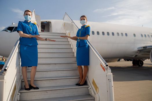 Full length shot of two beautiful air stewardesses in blue uniform and protective face masks looking at camera, standing on airstair and welcoming passengers. Aircrew, occupation, covid19 concept