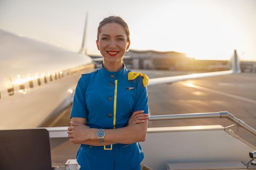 Portrait of beautiful air stewardess with red lips in blue uniform smiling at camera, posing outdoors with commercial airplane near the terminal in an airport in the background. Aircrew concept