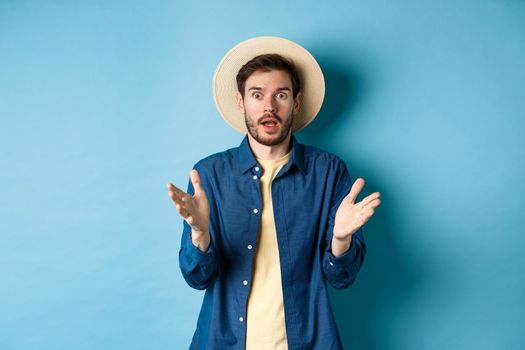 Shocked male tourist on vacation raising hands up and looking at camera startled, react to big news with disbelief, standing on blue background in straw summer hat and shirt.