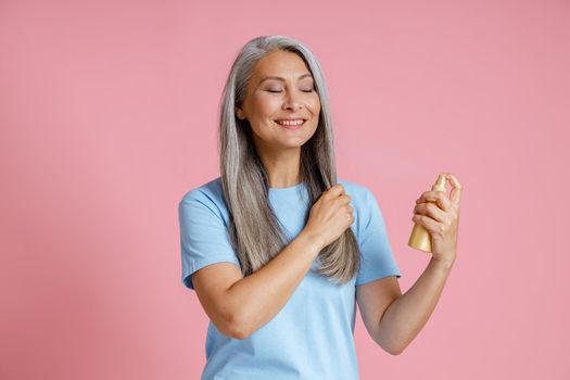 Positive Asian lady applies spray product onto loose grey hair standing on pink background in studio. Mature beauty lifestyle